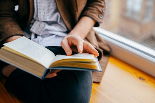 A person holds a book open to read while sitting on the window ledge