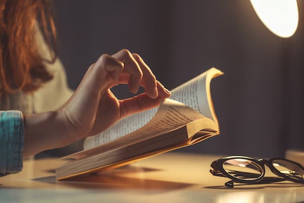 A person leafs through a book in low light. A pair of glasses sit on the table.