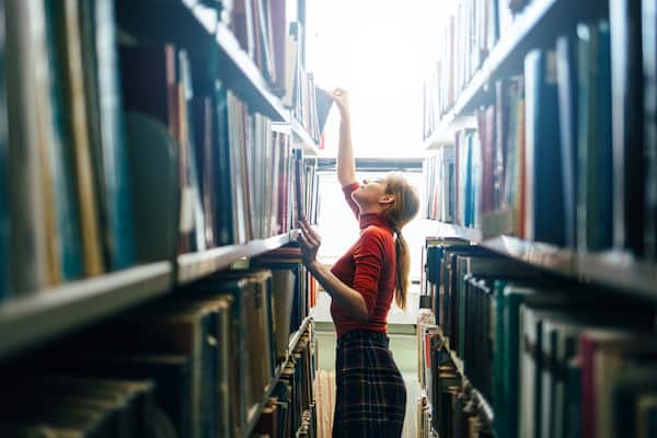 A person stands between shelves in a library and reaches for a reference book.