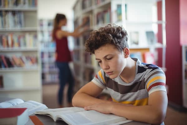 A young teenager places their arms across an open book and reads at a desk in a library.