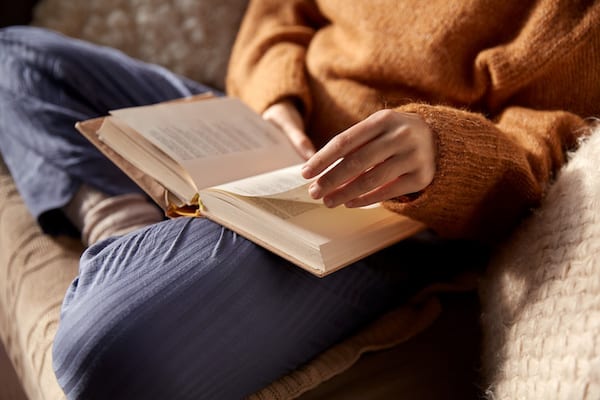 Someone sits reading cross-legged, wearing comfortable clothes on a sofa.