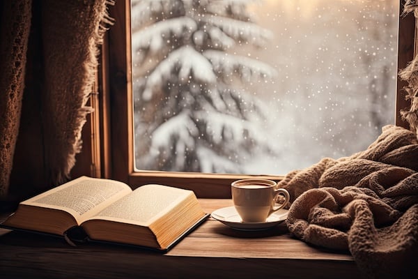 A wintry scene with a book, hot drink, and blanket in front of a snowy window