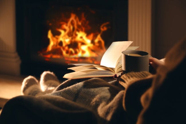 A person reading by the fire with a hot drink and a blanket