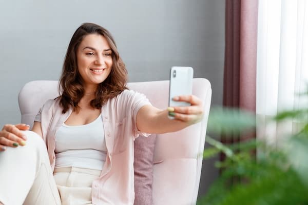 A person takes a selfie on their smartphone while sitting in a chair.