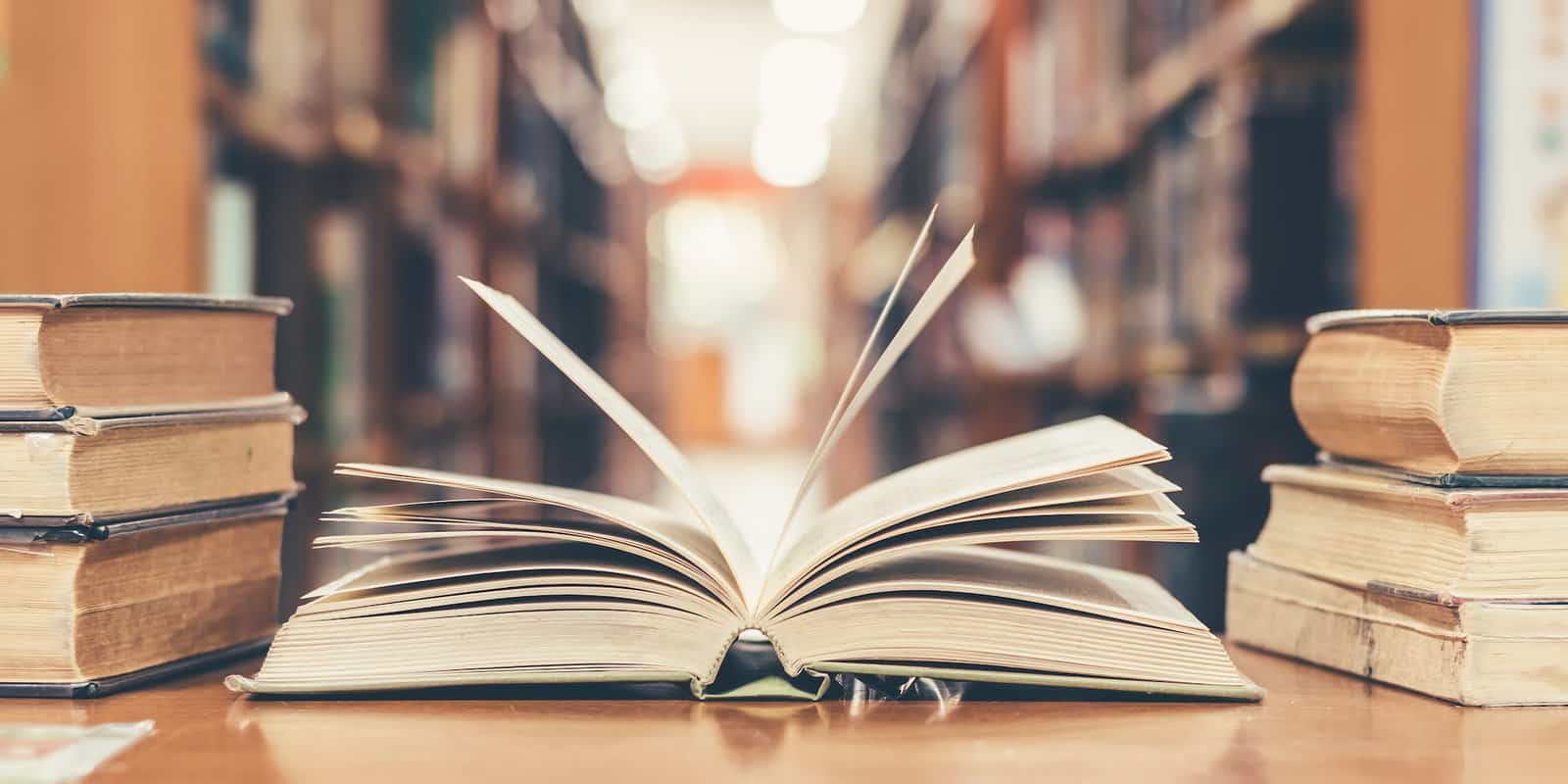 A book open next to two piles of books, on top of a desk within a library.