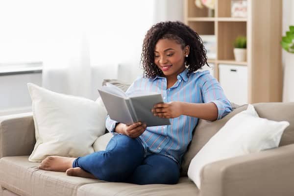 A young adult sitting on a sofa and reading a book in their living room.