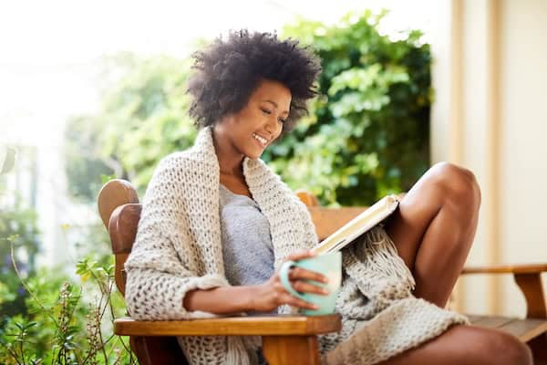A woman reading a book in a chair outside in the sun with a mug.