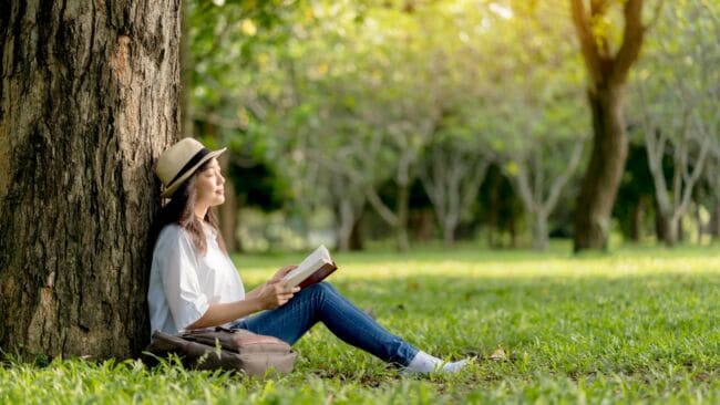 A woman reading a book, leaning against a tree on a sunny day.