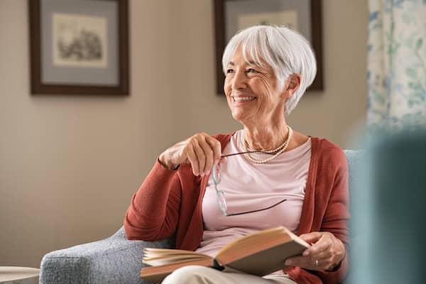 An older woman sitting in a chair holding her reading glasses as she smiles to herself with a book in hand.