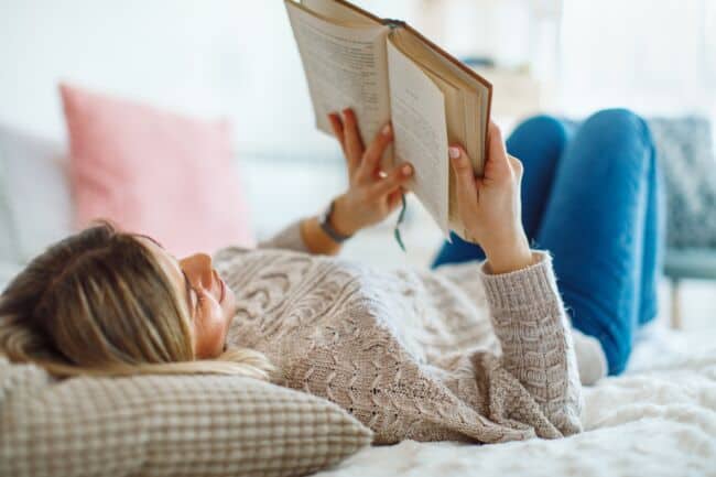 A woman lying back with her head on a cushion as she enjoys reading a book.