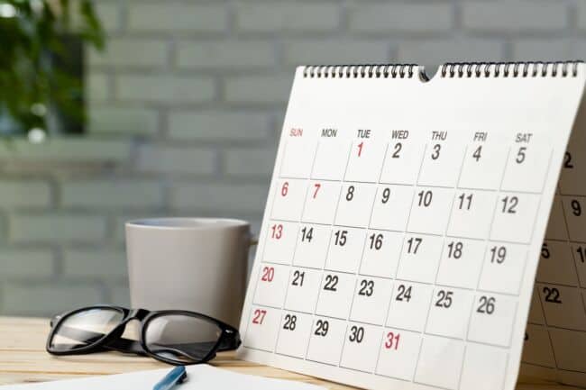 A calendar on a desk with a mug, pen, and a pair of glasses.