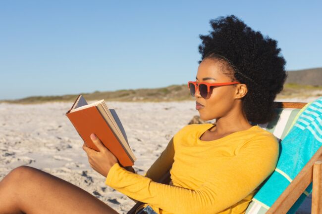 Woman sat on a deckchair engrossed in her book as she sits on the beach in summer.