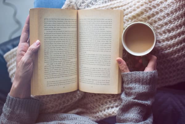 A person's hands resting on a blanket while holding a book and a cup of coffee.