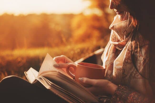 Alt text: A woman sits near a tree in an autumn park and holds a book and a cup with a hot drink in her hands.