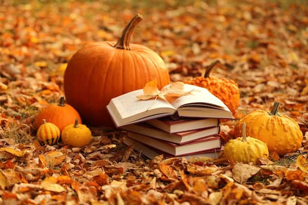 Alt text: Stack of books and orange pumpkins set on autumn foliage on blurred nature background.