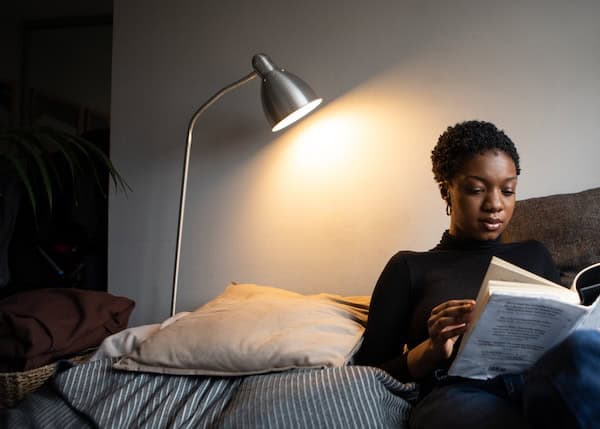 A woman sitting on the sofa reading lit by the light of a lamp.