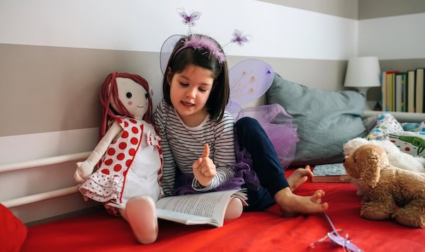 Little girl dressed as a butterfly sitting on the bed reading a book to her doll.